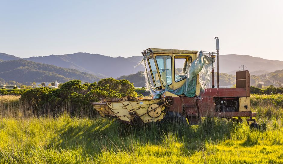 A yellow excavator with the branding 'dusseldorp' on its arm is positioned on a construction site, lifting a large black container or box. The machine is surrounded by loose soil, rocks, and construct