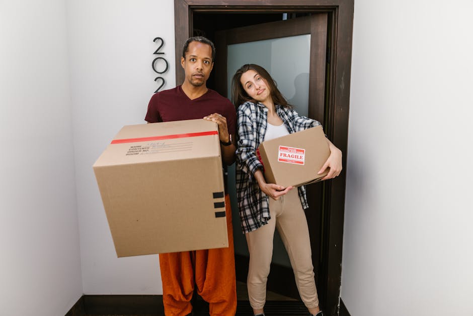 Two movers from Man with Van Aperfield are standing inside a narrow hallway in front of a doorway, each holding cardboard boxes filled with household items. The male mover, on the left, is wearing a maroon T-shirt and orange work trousers, while the female mover, on the right, is dressed in a checkered shirt and light-colored pants. The boxes are secured with packing tape; one has a label indicating it is fragile. Behind them is a plain white wall with the house number '200' visible, and the doorframe is dark wood. The environment appears well-lit, suggesting natural or bright indoor lighting, and the scene captures the process of home relocation and furniture transport as part of packing and moving activities. Man with Van Aperfield specializes in removals, including loading and transportation of household belongings, aligned with the context of house moving and logistics.