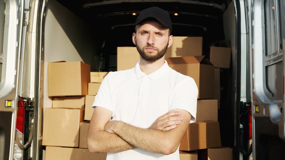 A man with a serious expression, wearing a black cap and a white polo shirt, stands inside the loading area of a moving van with the rear doors open. Behind him are numerous cardboard boxes of various