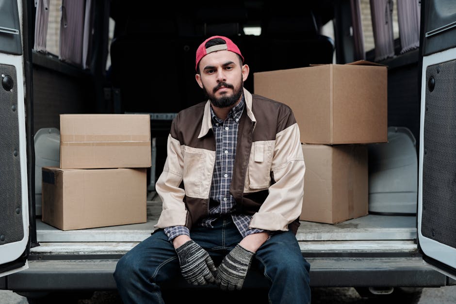 A man with a serious expression, wearing a black cap and a white polo shirt, stands inside the loading area of a moving van with the rear doors open. Behind him are numerous cardboard boxes of various