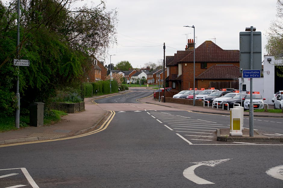 A street scene in Aperfield showing a residential area with a paved road and a pavement on the left side, lined with green trees and bushes. In the foreground, there is a loading zone with white painted lines and a small yellow and white barrier. A sign indicating 'Unsuitable for HGVs' is mounted on a pole near the loading area. To the right, there is a parking lot filled with white, silver, and gray cars parked alongside a multi-story brick building with chimneys and windows. The background features additional houses with pitched roofs, some with chimneys, and a cloudy sky overhead. The scene appears to be part of a home relocation or moving process, potentially involving a parking or loading permit from Bromley Council, as referenced on the page. The environment is calm with no visible activity, highlighting the street's suitability for furniture transport and house removals facilitated by Man with Van Aperfield.