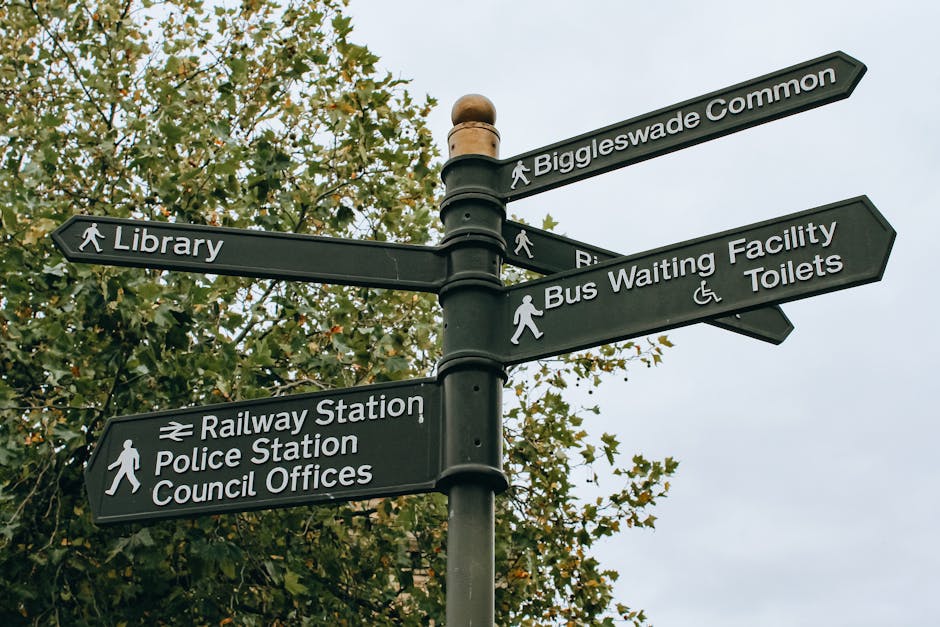 A black multi-directional street signpost with white text and icons, situated on a sidewalk against a backdrop of green leafy trees and an overcast sky. The signs indicate directions to various local amenities and facilities, including Biggleswade Common, Library, Railways Station, Police Station, Council Offices, Bus Waiting Facility, and Toilets. The signs are mounted on a single pole with windings, with pedestrian icons alongside each location name to signify walking directions. The surrounding environment appears to be in a rural or suburban area, with no vehicles or people visible in the immediate vicinity. This scene relates to local area navigation, which may be relevant in a context of house removals or community relocation services, offered by Man with Van Aperfield, to aid residents in planning their move or understanding local infrastructure for street loading permits.