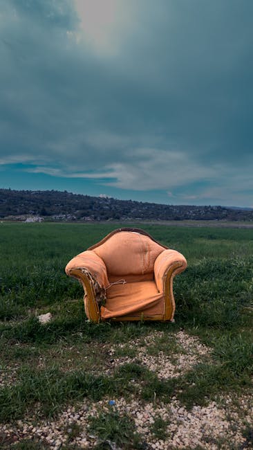 A worn, orange upholstered armchair with wooden detailing on the arms and legs, positioned on a patch of uneven grassy terrain outdoors. The fabric cover shows signs of damage and deterioration. The chair is situated in an open field with green grass extending into the distance, under a cloudy sky with patches of blue. In the background, there is a distant hillside and a cloudy sky, indicating an outdoor setting. The image appears to capture a scene of furniture disposal or an outdoor staging area related to home relocation or furniture transport, consistent with services offered by Man with Van Aperfield, as highlighted in their removals and bulky furniture disposal services.