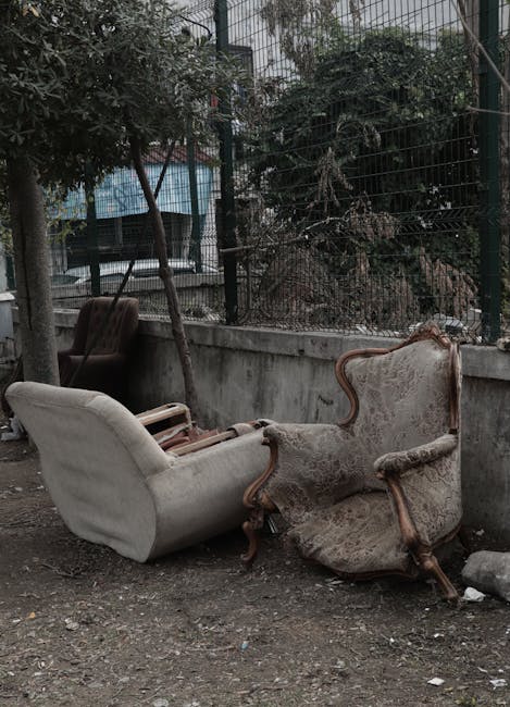 A vintage wooden sofa with patterned upholstery positioned on a pavement in front of a large pile of mixed waste paper, cardboard boxes, and packaging materials stacked against a wall. The waste includes crumpled newspapers, flattened boxes, plastic wrappers, and packaging inserts, some in bright colors like red, blue, and pink, while others are in brown paper or cardboard. The sofa appears to be part of a garbage disposal or bulky furniture removal process as seen in an urban setting, with nearby cardboard boxes and plastic waste scattered around. The scene is outdoors with visible natural lighting, illustrating a bulky furniture disposal situation where old furniture and household waste are being prepared for removal or recycling, aligning with home relocation and furniture transport services offered by Man with Van Aperfield.