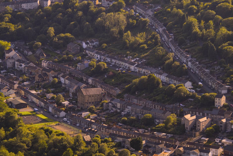 An aerial view of a residential area with rows of terraced houses and semi-detached homes, set among lush green trees and vegetation. The image shows a variety of buildings, including a large, historic stone structure with a pitched roof and exposed stone walls, likely part of a community or church. Surrounding the houses are paved streets, some with parked cars. In the foreground, a sports court with a green playing surface and boundary markings is visible, adjacent to gardens and backyards with lawn areas and fences. The scene is bathed in warm, natural light, highlighting the detail of the rooftops, trees, and the overall layout of the neighbourhood. This setting provides a context of a peaceful suburban environment, suitable for house removals and furniture transport, with Man with Van Aperfield involved in home relocation services that include packing, loading, and transporting belongings within the area.