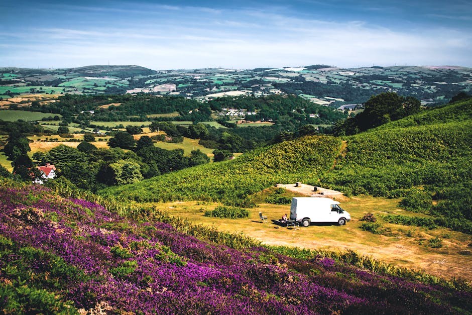 A white moving van parked on a grassy clearing surrounded by rolling green hills and vibrant purple and green foliage in a rural landscape. The van is positioned in the middle ground, with the back doors open. Nearby, there are two folding chairs and a small table, suggesting a break during the furniture transport or home relocation process. The scene features distant fields, trees, and a patchwork of pastures, with a partly cloudy sky overhead providing natural light. The location appears to be in a countryside setting, consistent with the scenic views associated with house removals and relocation services, as offered by Man with Van Aperfield. The image captures a peaceful moment in a rural environment, emphasizing the logistical aspects of moving goods through outdoor environments and the preparation involved in packing and transporting personal belongings.