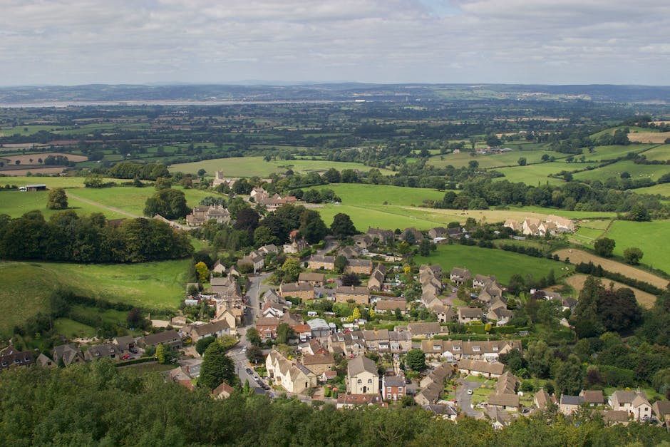 An aerial view of a residential area with rows of terraced houses and semi-detached homes, set among lush green trees and vegetation. The image shows a variety of buildings, including a large, historic stone structure with a pitched roof and exposed stone walls, likely part of a community or church. Surrounding the houses are paved streets, some with parked cars. In the foreground, a sports court with a green playing surface and boundary markings is visible, adjacent to gardens and backyards with lawn areas and fences. The scene is bathed in warm, natural light, highlighting the detail of the rooftops, trees, and the overall layout of the neighbourhood. This setting provides a context of a peaceful suburban environment, suitable for house removals and furniture transport, with Man with Van Aperfield involved in home relocation services that include packing, loading, and transporting belongings within the area.