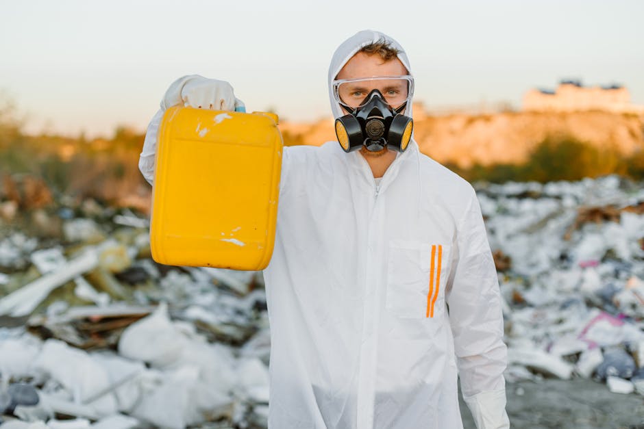 A person wearing a white protective coverall, safety goggles, and a black respirator mask stands outdoors in front of a large pile of mixed household waste and debris, including plastic, cardboard, and other refuse, on a landfill site. The individual is holding a yellow container, which appears to be used for hazardous waste collection, with their right hand. The background features a landscape with a lightly cloudy sky and distant structures bathed in warm sunlight, highlighting the scene of waste management and hazardous waste disposal. This visual relates to house removal services where careful handling and proper disposal of waste, especially hazardous materials, are essential, as outlined in the Hazardous Waste Rules for Aperfield residents, explained by Man with Van Aperfield.