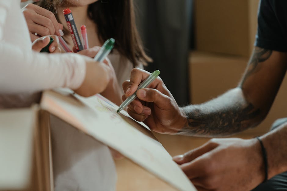 Close-up view of a person's hand using a green marker to label cardboard boxes and packaging materials for home relocation, with additional hands seen holding pens of different colours, possibly marking or packing items. The boxes are arranged on a wooden surface inside a property, indicating a packing and moving process. In the background, blurred figures of more people working together are visible, highlighting teamwork during a house move. The environment appears well-lit with natural light, and the scene illustrates the preparatory stages of furniture transport and packing for a home removal. This visual aligns with the services offered by Man with Van Aperfield, supporting logistics and packing for tight access homes on Aperfield Lane.