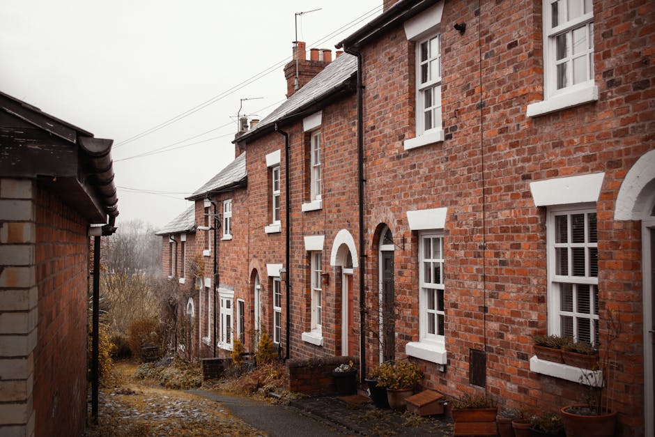 View from the top of a staircase inside a home showing two women carrying cardboard moving boxes down the steps, while a third woman waits at the bottom for assistance. The staircase has black metal railings, and the walls are painted white with a warm light fixture mounted on one side. A small potted plant on a ledge near a window is visible at the top of the stairs, providing natural light. The cardboard boxes are sealed with packing tape, and one has a framed photograph placed on top. The scene captures the process of home relocation, with individuals engaged in furniture transport and packing and moving activities, supported by manual handling equipment such as trolleys or straps used during loading and unloading. Man with Van Aperfield are involved in the logistics of this furniture transport, focusing on tight access and staircase navigation as part of their removals service.