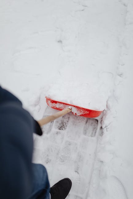 A person in dark clothing and black footwear is using a red snow shovel to clear snow from a pavement outside a building. The shovel is partially submerged in the snow, which appears thick and fresh.