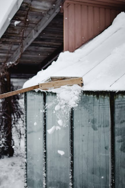 A person in dark clothing and black footwear is using a red snow shovel to clear snow from a pavement outside a building. The shovel is partially submerged in the snow, which appears thick and fresh.
