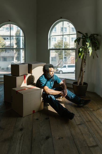 A man with curly hair and dark skin, wearing a blue t-shirt and jeans, sits on the wooden floor of a room with large, arched windows allowing natural daylight to illuminate the space. He is surrounded
