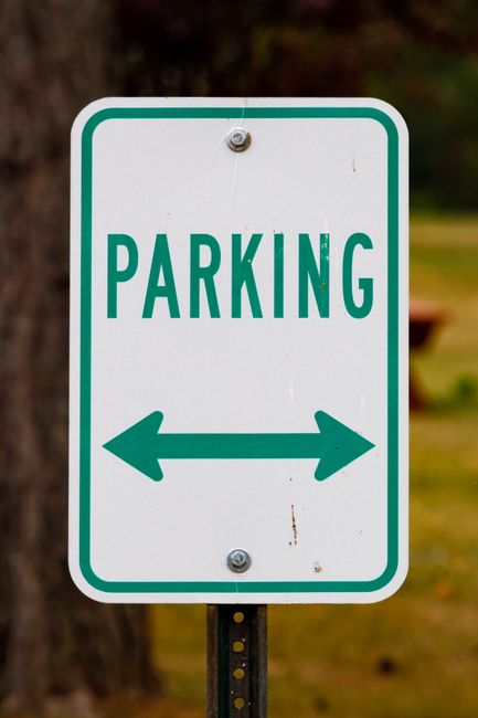 A large white directional arrow painted on an asphalt surface, pointing downwards and slightly to the right, within a rectangular box outlined in white. The surface appears to be part of a parking are