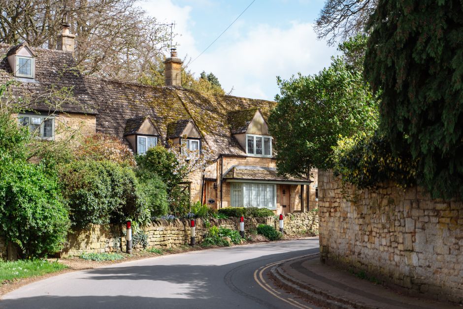 A residential street in Aperfield featuring a traditional stone house with a sloped roof, dormer windows, and a chimney, situated behind a low stone wall with lush greenery and trees on both sides. The house is partially obscured by leafed trees and shrubs, with the front garden visible in front of the house. The pavement curves gently to the right, and there are white and red reflective traffic bollards along the edge of the low stone wall. The scene is well-lit with natural daytime light, and a clear sky with some clouds overhead. This setting is relevant for house removal services, illustrating the type of period home where careful furniture transport and packing are necessary during a home relocation, as managed by companies like Man with Van Aperfield.