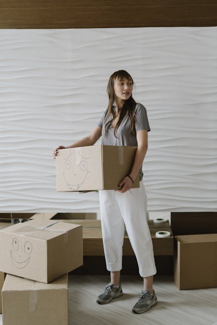A young woman with long brown hair, dressed in a grey t-shirt and white trousers, stands indoors on a light wooden floor holding a cardboard box with a smiley face drawn on the side. She appears to be in the process of packing or preparing for a home relocation, with additional cardboard boxes nearby—one on the floor and another partially open on a ledge. The background features a textured white wall with a wavy pattern, and a bench with a few small objects or perhaps rolled-up documents placed on it. The scene is well-lit with natural or soft artificial light, suggesting a professional or domestic setting involved in packing and moving activities, which aligns with the services offered by Man with Van Aperfield as part of their house removals and furniture transport operations.