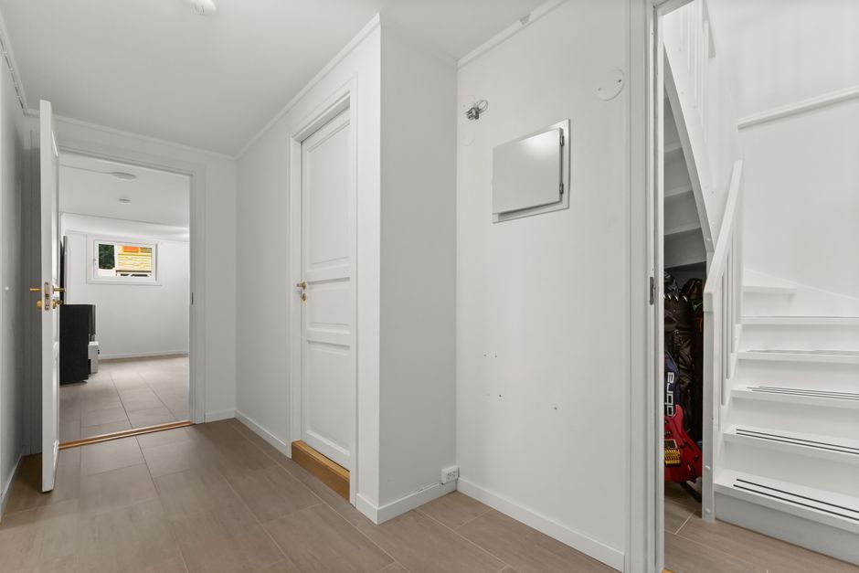 Inside a residential property, the image shows a narrow hallway with white walls and wooden flooring, leading to a staircase with white-painted steps and a matching railing, positioned on the right side of the scene. To the left of the staircase, a storage closet or utility cupboard with a white door is partially open, revealing various tools and bags inside. Opposite the staircase, there is a small access panel or electrical box mounted on the wall. In the background, a doorway opens into a room with a window, allowing natural light to illuminate the space, and a black piece of furniture is visible in that room. The environment appears clean and tidy, with professional moving equipment such as furniture blankets or straps not visible in this image but typically used during home relocation. This setting is consistent with a scene where [COMPANY_NAME] assists with staircase and narrow hall moves within homes or flats, focusing on safe and efficient furniture transport during packing and moving processes for property relocations in Aperfield.
