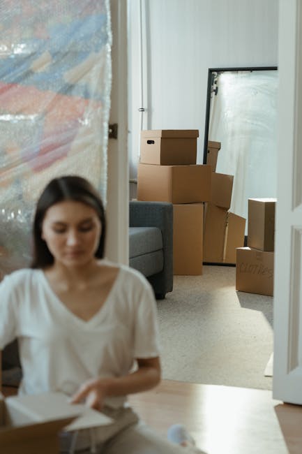 Inside a residential property, a young woman with long dark hair, dressed in a white top, is seated on a wooden floor, focusing on packing or organizing items in a cardboard box. In the background, there are several stacked and scattered cardboard boxes of various sizes, some open, indicating a packing or move-in process. A grey upholstered sofa is partially visible, positioned along the wall, with a large mirror propped against the wall behind the boxes. The room is well-lit with natural light coming through a window or door, and the environment appears to be a staging or staging-ready space during home relocation. This scene depicts the packing and moving logistics involved in furniture transport and home relocation services, as managed by Man with Van Aperfield, in a typical apartment or flat setting in Aperfield as referenced on the company’s website.