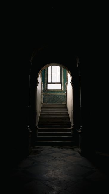 Inside a residential property, the image shows a narrow hallway with white walls and wooden flooring, leading to a staircase with white-painted steps and a matching railing, positioned on the right side of the scene. To the left of the staircase, a storage closet or utility cupboard with a white door is partially open, revealing various tools and bags inside. Opposite the staircase, there is a small access panel or electrical box mounted on the wall. In the background, a doorway opens into a room with a window, allowing natural light to illuminate the space, and a black piece of furniture is visible in that room. The environment appears clean and tidy, with professional moving equipment such as furniture blankets or straps not visible in this image but typically used during home relocation. This setting is consistent with a scene where [COMPANY_NAME] assists with staircase and narrow hall moves within homes or flats, focusing on safe and efficient furniture transport during packing and moving processes for property relocations in Aperfield.